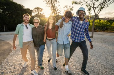 Gün batımında parka doğru yürürken ve birlikte gülerken kollarını birbirlerine dolamış mutlu ve çeşitli genç arkadaşlardan oluşan uzun bir çekim..