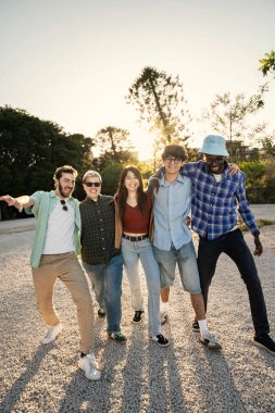 Çok ırklı bir grup genç arkadaş, kollarını birbirlerine dolamış gün batımında bir yolda birlikte yürüyorlar. Tam boy dikey çekim arkadaşlığı ve ortak yolculuğu temsil eder..