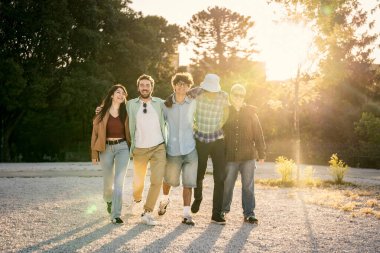 Birbirlerine sarılmış, gün batımında bir yolda birlikte yürüyen, uzun gölgeler yaratan çok ırklı bir arkadaş grubu. Arkadaşlık, birlik ve ilerleme kavramı.