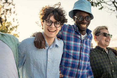 Neşeli ve çeşitli etnik çeşitlilikteki yetişkin arkadaşlar kameraya bakar ve neşeli bir şekilde gülerler. Güneşli bir günde, arka planda güzel bir ışık olan bir parkta birlikte eğlenirler..
