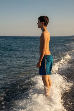 Huzurlu bir genç, sahilde güzel bir yaz günbatımında, denizde dururken, ferahlatıcı bir dalga ayaklarına vururken, gözleri kapalı farkındalık pratiği yapıyor.