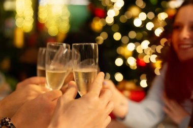 Arkadaşların ellerinin kadehte şampanya kadehlerini tutarken, bir Noel ağacının bulanık arkaplanı sıcak bir altın bokeh etkisi yaratıyor..