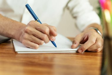 Mavi bir kalem tutan ve çizgili bir not defterine yazı yazan bir adamın, toplantı sırasında not aldığı ya da kontrat imzaladığı detaylı bir yakın çekim..