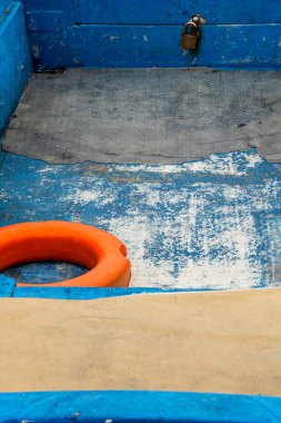 Eski bir balıkçı teknesinin yıpranmış ve soyulmuş mavi zemininin soyut yüksek açılı görüntüsü, parlak turuncu bir can simidi, güçlü bir kavramsal renk karşıtlığı oluşturuyor..