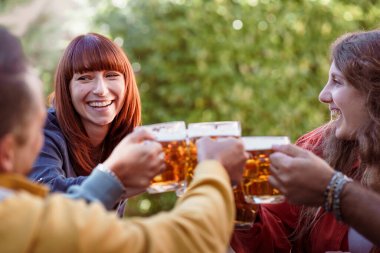 Bir grup mutlu genç arkadaş açık hava bahçe partisinde bira bardaklarıyla gülüp kadeh kaldırıyor. Arkadaşına gülümseyen neşeli bir kadına odaklan. Otantik bir bağlantı anını yakala..