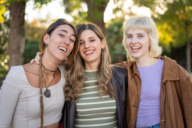 Üç farklı genç kadından oluşan coşkulu bir grup, en iyi arkadaşlar, altın saatin sıcacık ışığı altında bir parkta kollarıyla mutlu bir şekilde gülüyorlar. Otantik arkadaşlık, neşe ve gençlik kültürü kavramı..