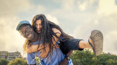 Mutlu, çok kültürlü bir çift güneşli bir yaz gününde dışarıda eğleniyor. Gülümseyen genç kadın, erkeğe arkadan sarılıyor, çeşitlilik konsepti, arkadaşlık, aşk, neşe, kaygısız yaşam tarzı, seyahat ve tatil.