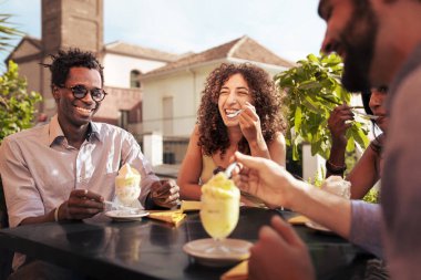 Neşeli, çok kültürlü bir grup genç arkadaş, İtalya 'nın Sicilya kentinde güneşli bir yaz tatili sırasında bir açık hava kafede geleneksel granita tatlısını yerken kahkahalarını paylaşıyor..