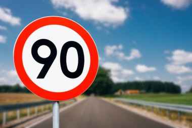 Arkasında ağaçlar olan hız limiti tabelası. Saatte maksimum 90 kilometre. Yol geçmişinde güvenlik var. Beyaz, yuvarlak, kırmızı sınır çizgisi. Trafik cezası geçmişi. Hız iyi..