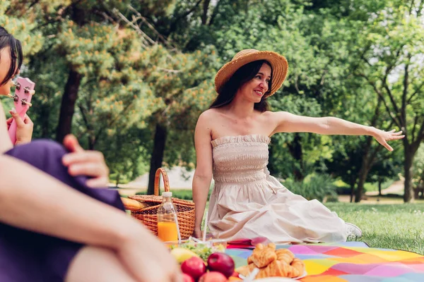 Happy young female in summer dress and straw hat sitting on blanket with crop friends on green lawn and enjoying picnic in park