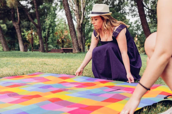 Young female in summer dress and hat placing colorful picnic blanket with the help of crop friend on meadow in park