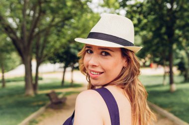 Glad female in hat with long hair smiling and looking at camera over shoulder on path on summer day in park