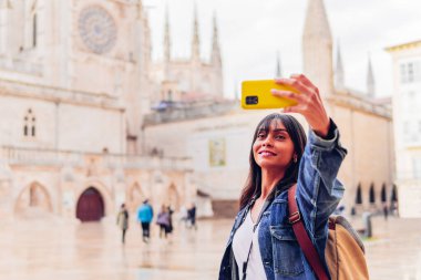 Young Hispanic lady in denim jacket with backpack using phone to take selfie against Burgos Cathedral in Castile and Leon