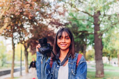 Cheerful young Hispanic female in denim jacket and with backpack holding photo camera and smiling at camera while standing in city park