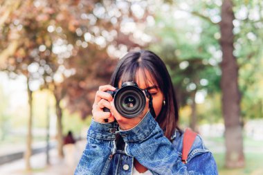 Unrecognizable female photographer in denim jacket taking picture on photo camera while standing in green park in daylight