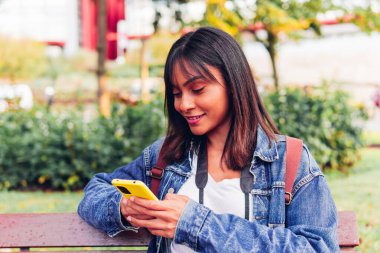 Young smiling female in denim jacket with backpack and photo camera browsing mobile phone while sitting on wooden bench near green bushes