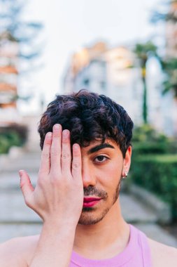 Siyahi kıvırcık saçlı, pembe dudaklı, kendine güveni tam genç bir erkek. Kameraya bakıp bulanık sokakta dikiliyor.