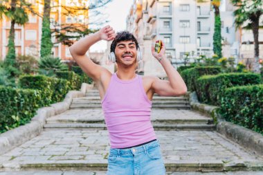 Pembe tişörtlü, kot pantolonlu, mutlu, genç bir erkek. Kablosuz kulaklıklı, akıllı telefonlu müzik dinlerken ve kollarını kaldırmış sokakta dans ederken gülümsüyor.