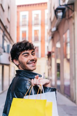 Deri ceketli, pozitif, genç, İspanyol bir erkek. Alışveriş torbalarıyla sokakta yürürken gülümsüyor ve kameraya bakıyor.