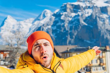 Sıcak elbiseli ve şapkalı hayret verici erkek turist heyecanla ağzını açıyor ve İsviçre 'de güneş ışığında kayalık dağlara karşı kendi portresini çekerken tatil köyünü işaret ediyor.