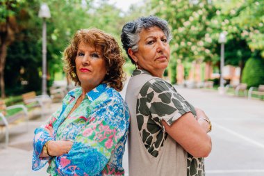 Ciddi olgun kadınlar kameraya bakıyor ve parkta kollarını kavuşturup poz veriyorlar.