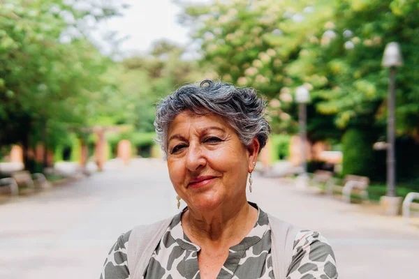 Positive middle age female in casual clothes with earrings smiling and looking at camera while standing on asphalt street beside blurred green trees park in daylight