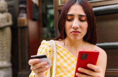 Sokakta oturan, kredi kartı ve cep telefonuyla ödeme yapmakta zorlanan endişeli bir kadının yakın plan fotoğrafı.