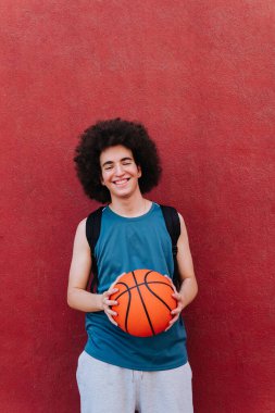 Kırmızı duvarın yanında topu olan genç bir basketbolcunun dikey portresi.