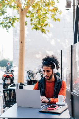 Laptop ve kulaklık kullanan Latin bir adamın açık hava kafeteryasında otururken çekilmiş dikey fotoğrafı.