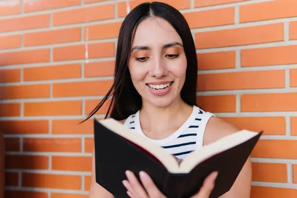 Smiling female student reading a book next to a brick wall in the campus