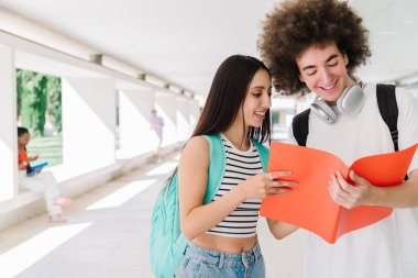 Sırt çantalı iki gülümseyen öğrenci üniversite koridorundaki bir klasörden notları inceliyor.