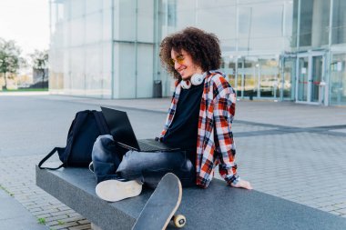 Modern bir üniversitede kaykay ve sırt çantasının yanında otururken dizüstü bilgisayar kullanan kıvırcık saçlı ve kulaklıklı genç bir adam.