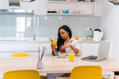 Genç bir kadın, taze meyve ve parlak dekorla dolu modern bir mutfakta akıllı telefonunu karıştırırken sağlıklı bir kahvaltıdan zevk alıyor.
