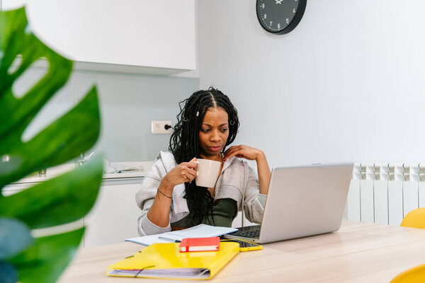Young woman working remotely from home drinking coffee while using laptop and sitting at kitchen table
