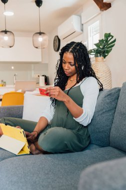 Hamile kadın evdeki koltukta oturuyor, dijital tablet ve kredi kartı kullanıyor, online alışveriş yapıyor.