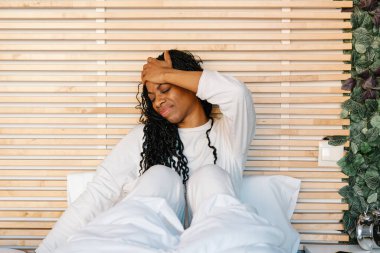 Genç bir kadın yatakta oturuyor, baş ağrısı çekiyor, sabah ışığıyla çevrili iken nazikçe alnını bastırıyor.