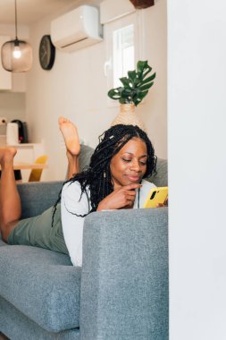 Huzurlu bir kadın boş zamanlarında rahat bir şekilde kanepede uzanıyor ve evinin rahat ortamında akıllı telefonunu kullanıyor.