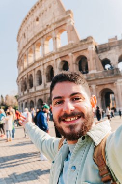 Genç sakallı turist, Roma, İtalya 'da kolezyumda kollarını açarak selfie çekiyor ve güneşli bir tatil gününün tadını çıkarıyor.