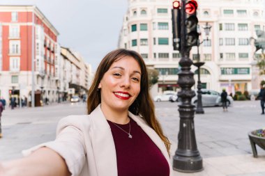 Genç iş kadını gülümsüyor ve şehir merkezinde kırmızı ışık ve bulanık şehir geçmişi olan bir sokakta selfie çekiyor.