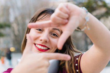 Neşeli genç bir kadın dışarıda gülümserken elleri ve parmaklarıyla bir çerçeve hareketi yapıyor.