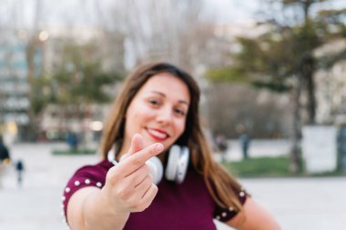 Bir şehir parkında boynuna kulaklık takmış, Kore kalbi sembolü yapan gülümseyen genç bir kadın.