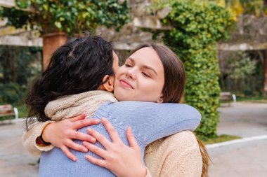 Parkta kucaklaşan iki neşeli kadın, samimi bir yakınlık anlarını paylaşıyorlar ve doğanın ortasında güçlü dostluklarını kutluyorlar.
