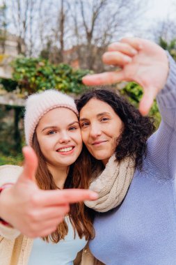 Gülümseyen anne ve kız elleriyle bir çerçeve hareketi yapıyor, parkta birlikte bir günün tadını çıkarıyorlar.