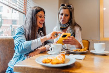 Bir kafede kahve ve kruvasan içen iki neşeli kadın, rahat bir sabahta sosyal medyada rahatça gezinirken kahkahalarını ve bağlantılarını paylaşıyorlar.