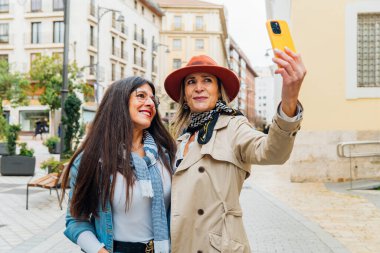 İki yetişkin kadın turist, şehir caddesinde yürürken akıllı telefonlarıyla selfie çekiyorlar. Birlikte tatillerinin tadını çıkarıyorlar ve anılarını yakalıyorlar.