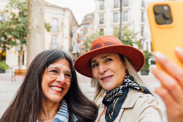 İki neşeli olgun kadın, biri şık bir şapka ve eşarp takıyor, resimli bir şehir meydanında akıllı bir telefonla selfie çekiyor, birlikte vakit geçiriyorlar.