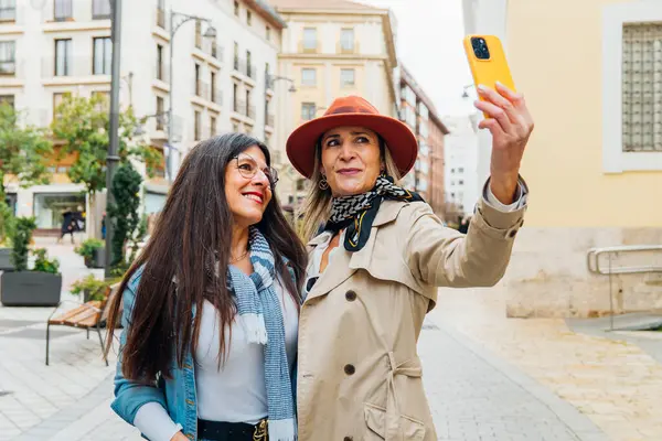 İki yetişkin kadın turist, şehir caddesinde yürürken akıllı telefonlarıyla selfie çekiyorlar. Birlikte tatillerinin tadını çıkarıyorlar ve anılarını yakalıyorlar.