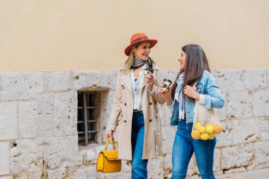 Sokakta yürüyen iki kadın, ellerinde tekrar kullanılabilir kahve fincanları ve içinde portakal ve limon bulunan bir alışveriş torbası, sürdürülebilir bir yaşam tarzı sergiliyorlar.
