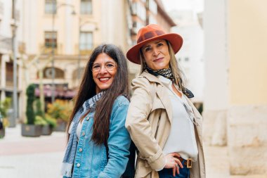 İki olgun, şık kadın, bir şehir caddesinde sırt sırta dururlar, güven ve dostluk yayarlar, kentsel bir çevrede kadın gücünü somutlaştırırlar.