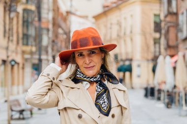 Zarif olgun kadın turuncu şapkasını düzeltirken büyüleyici bir Avrupa şehir meydanında geziniyor, kendine güveni ve sonsuz bir zarafeti var.